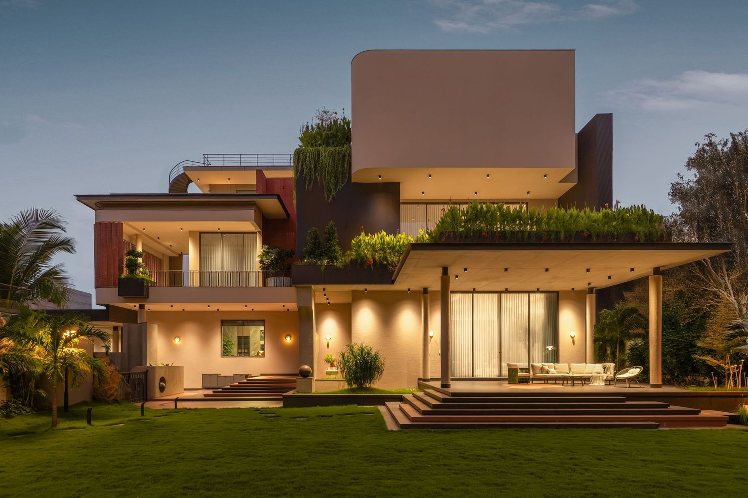 Modern luxury villa with green rooftop garden, illuminated facade, and manicured lawn at dusk.