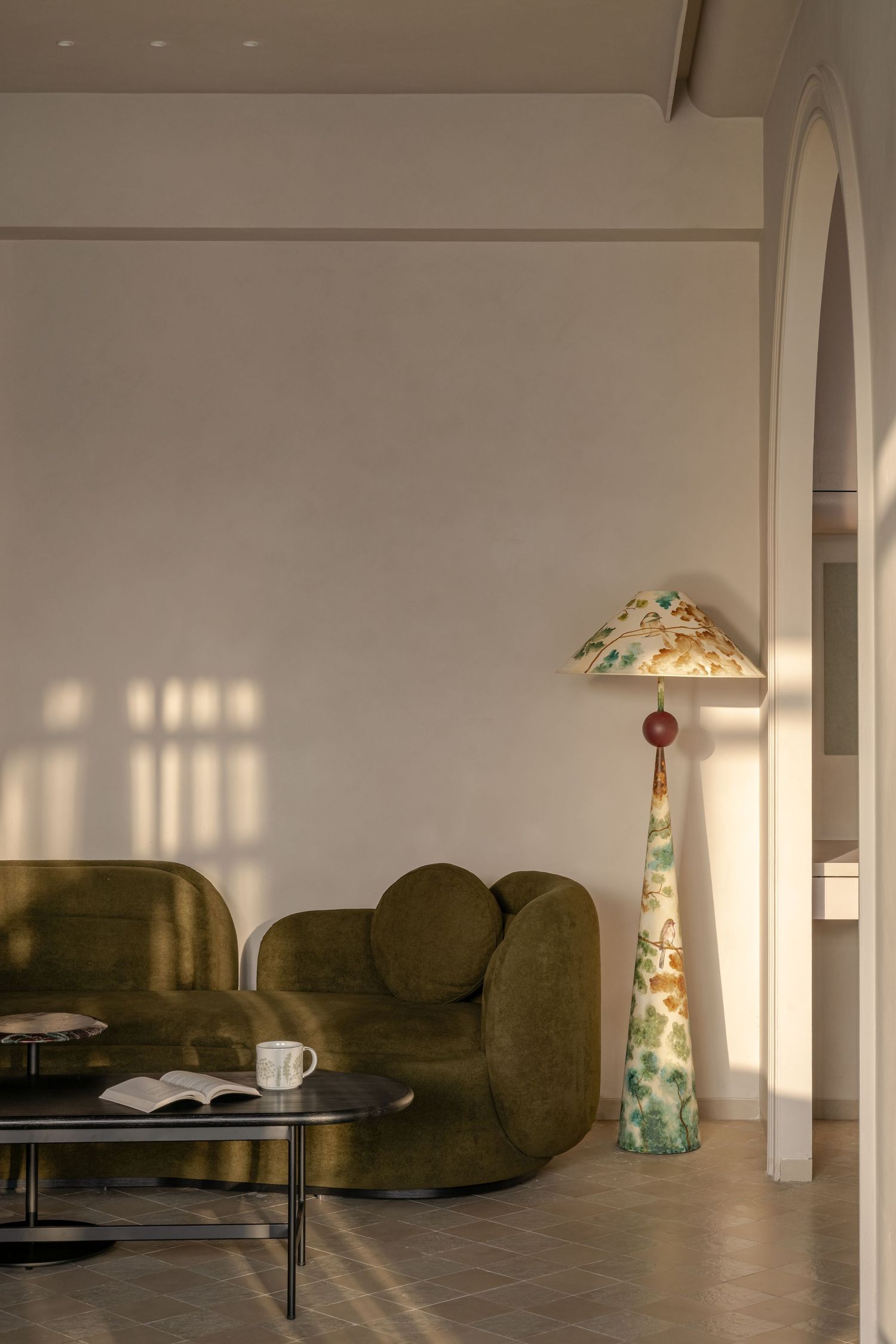 Warm sunlit living room with olive green curved sofa, black coffee table, and tall decorative ceramic floor lamp.