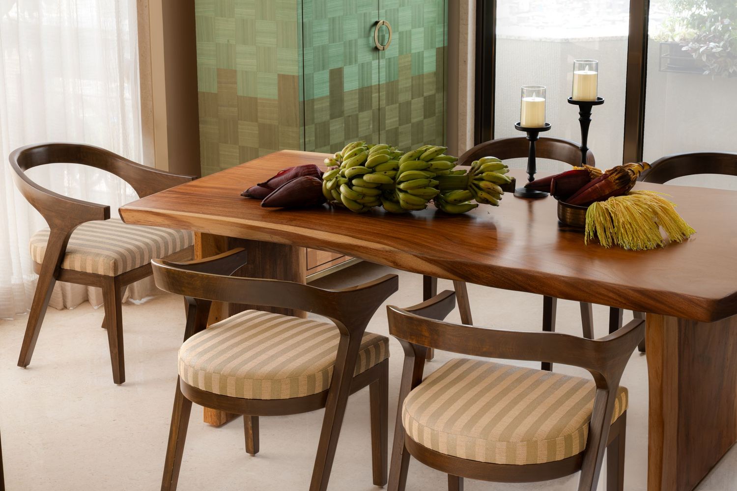 Rustic wooden dining table with live edge, striped cushion chairs, bananas centerpiece, and candles in bright room.