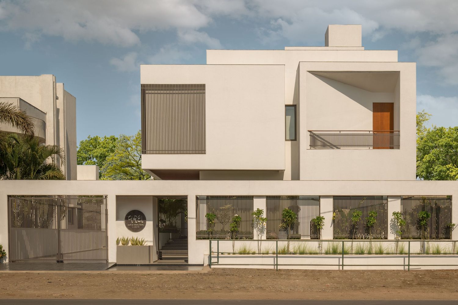 Modern white residential building with geometric facade, wooden gate, circular logo signage, and manicured hedges.