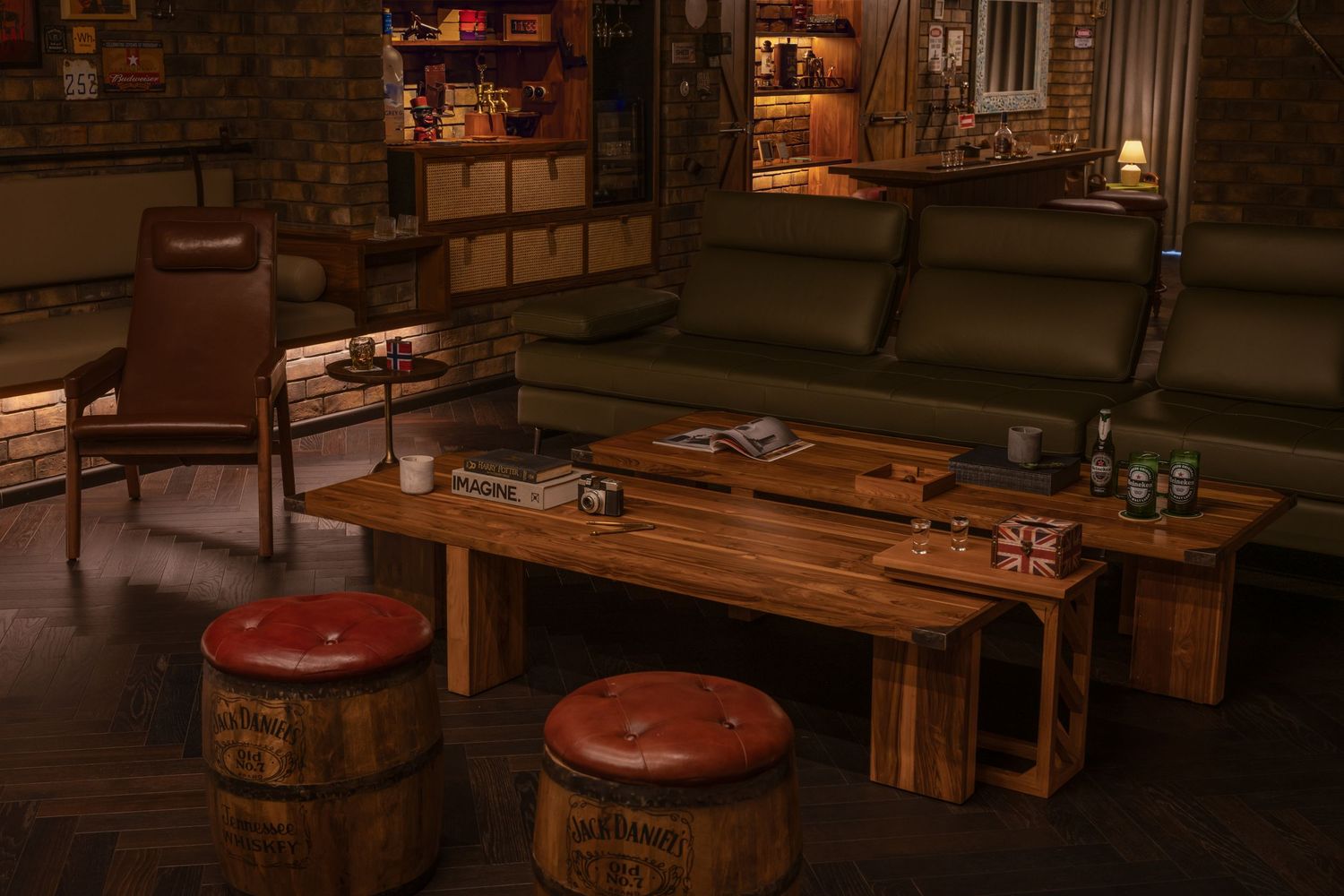 Dimly lit retro lounge featuring green leather sofa, wooden coffee table, barrel ottomans, and shelved bar backdrop.