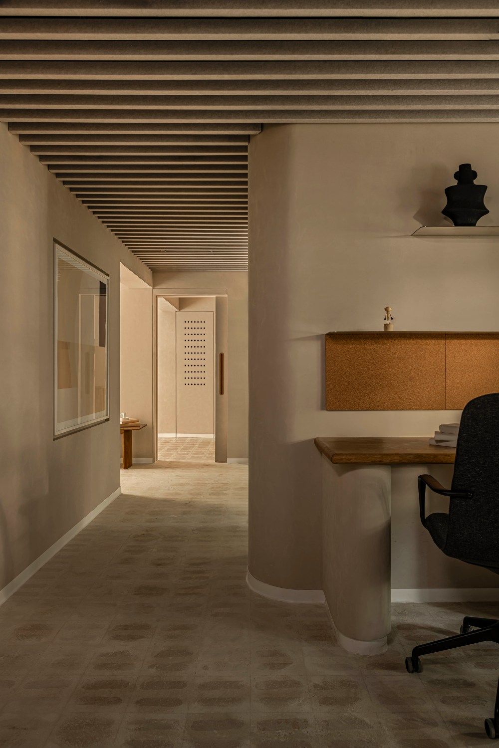 Dimly lit beige office corridor with slatted ceiling, wooden desk, and ergonomic chair in alcove.