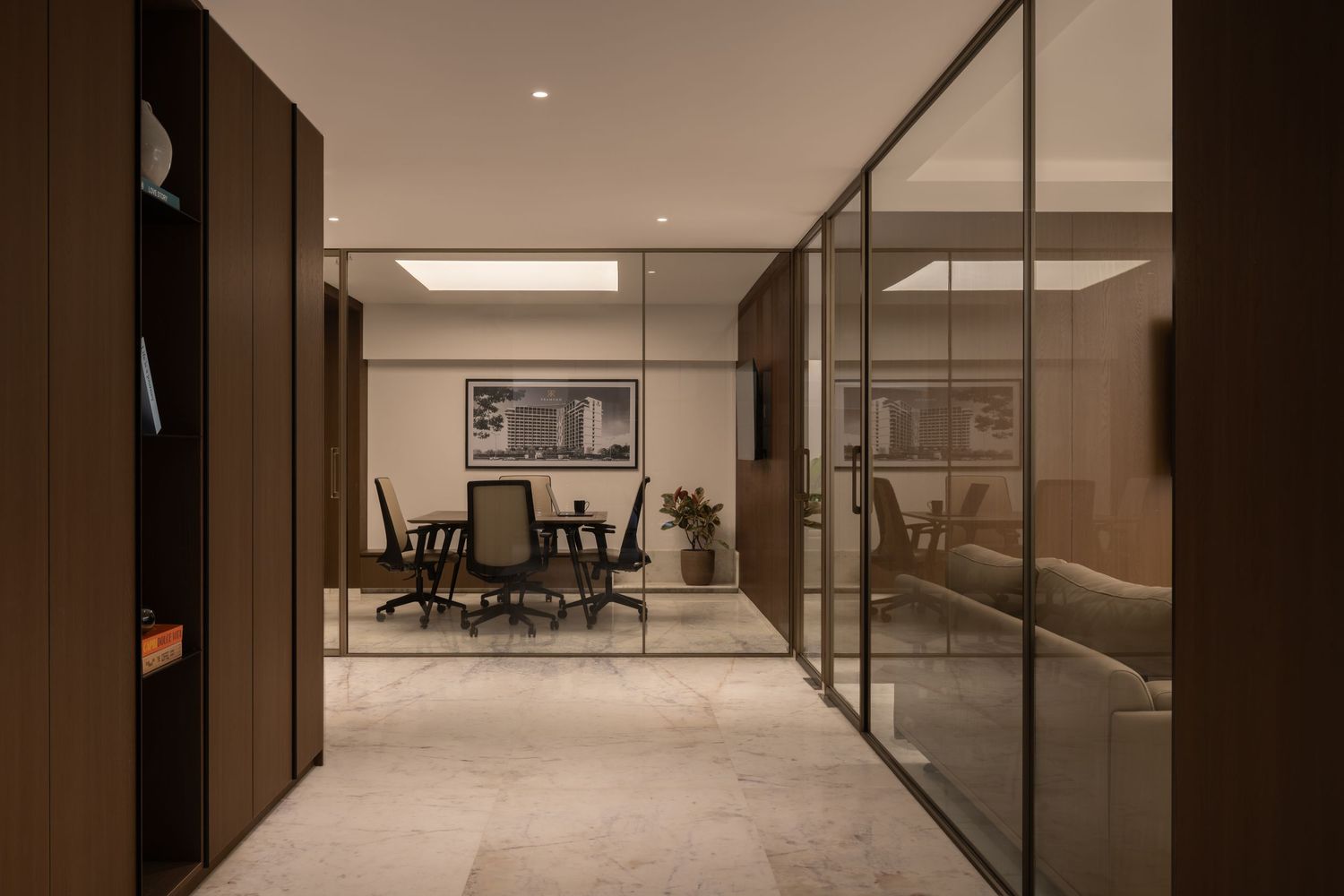 Sophisticated office interior with glass partition walls revealing a dark-furnished meeting room on marble flooring.