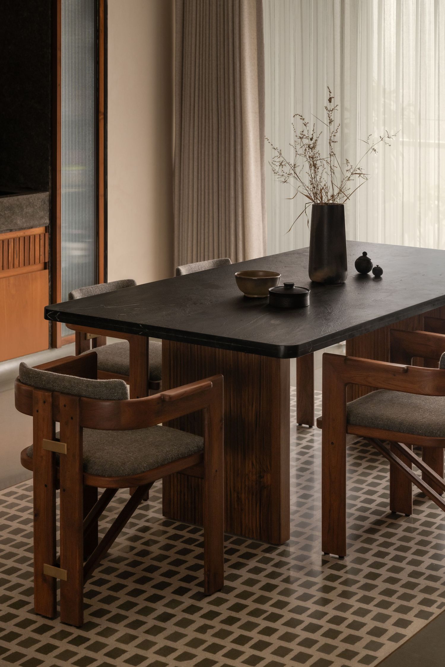 Minimalist dining space with black stone table, wooden chairs, dried floral centerpiece, and geometric patterned rug.