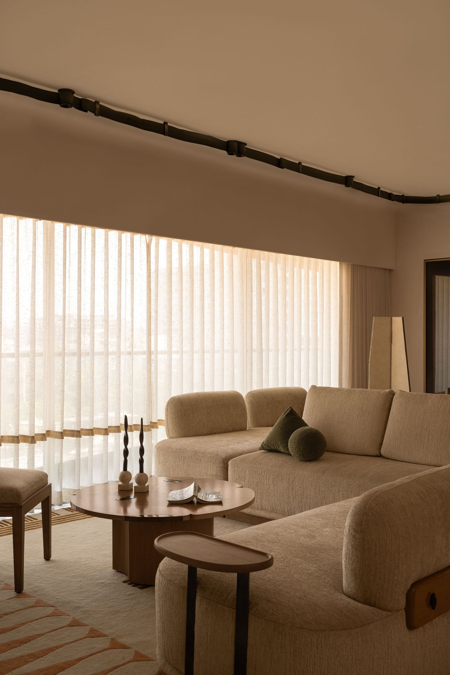 Warm beige living room with curved sofa, round wooden coffee table, and sheer vertical blinds.