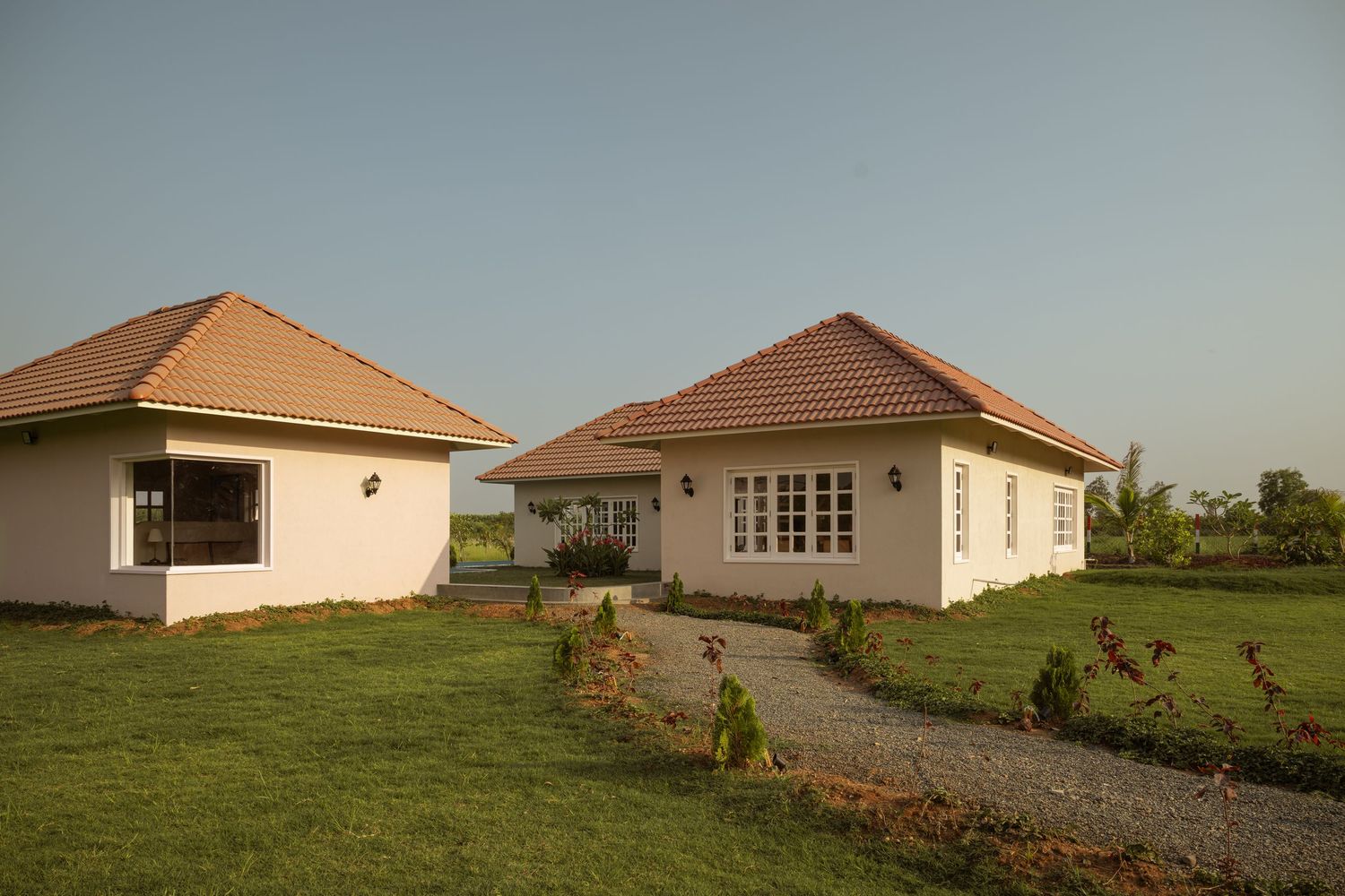 Two colonial bungalows with terracotta tile roofs, white arched doors, French windows, and lush green lawns at golden hour.