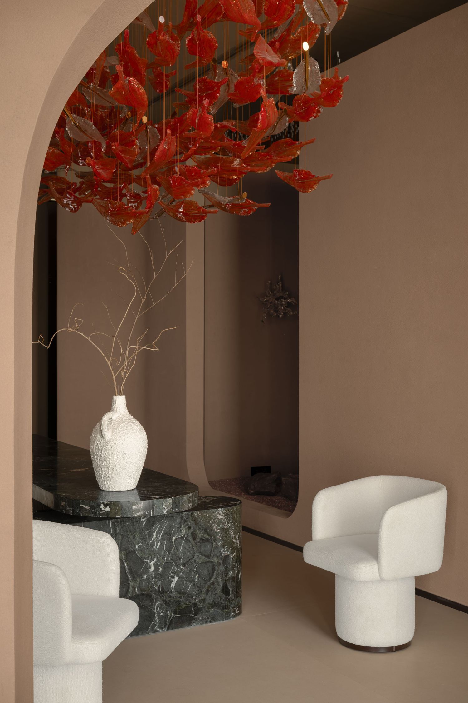 Modern interior with white armchairs, marble table, white ceramic vase, and red autumn leaf chandelier in arched alcove.