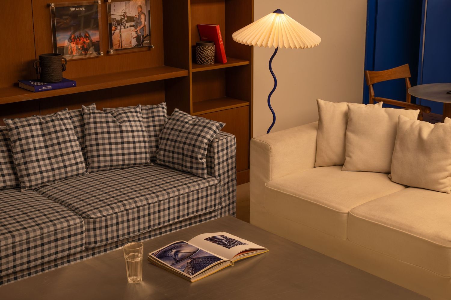 Cozy lounge with plaid sofa, white daybed, wavy blue floor lamp, and warm wooden shelving.