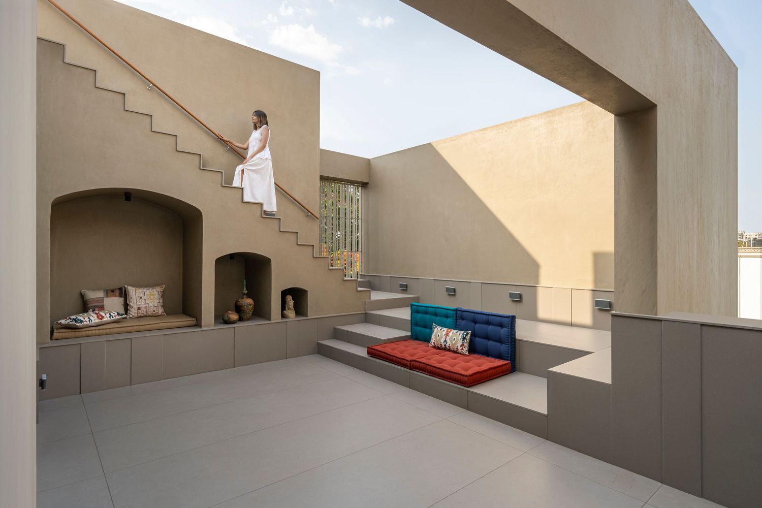 Modern minimalist courtyard with beige walls, built-in seating, colorful cushions, and a staircase with a person in white.