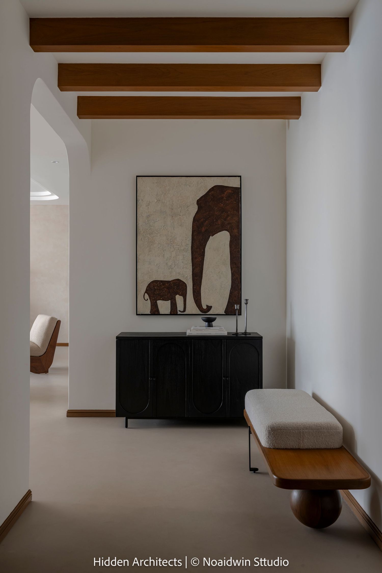 Minimalist hallway with wooden ceiling beams, large elephant artwork, black sideboard, and upholstered bench on neutral flooring.