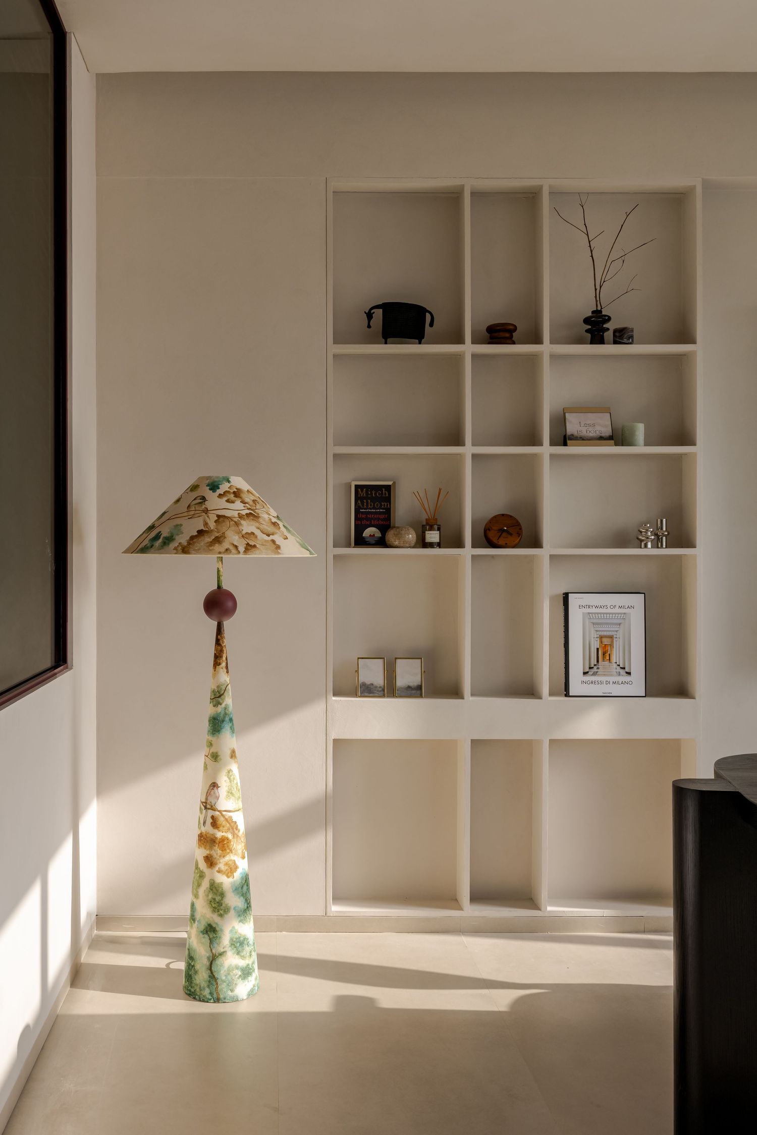 Cream built-in shelving unit with decorative objects beside a tall artistic ceramic floor lamp in warm sunlight.