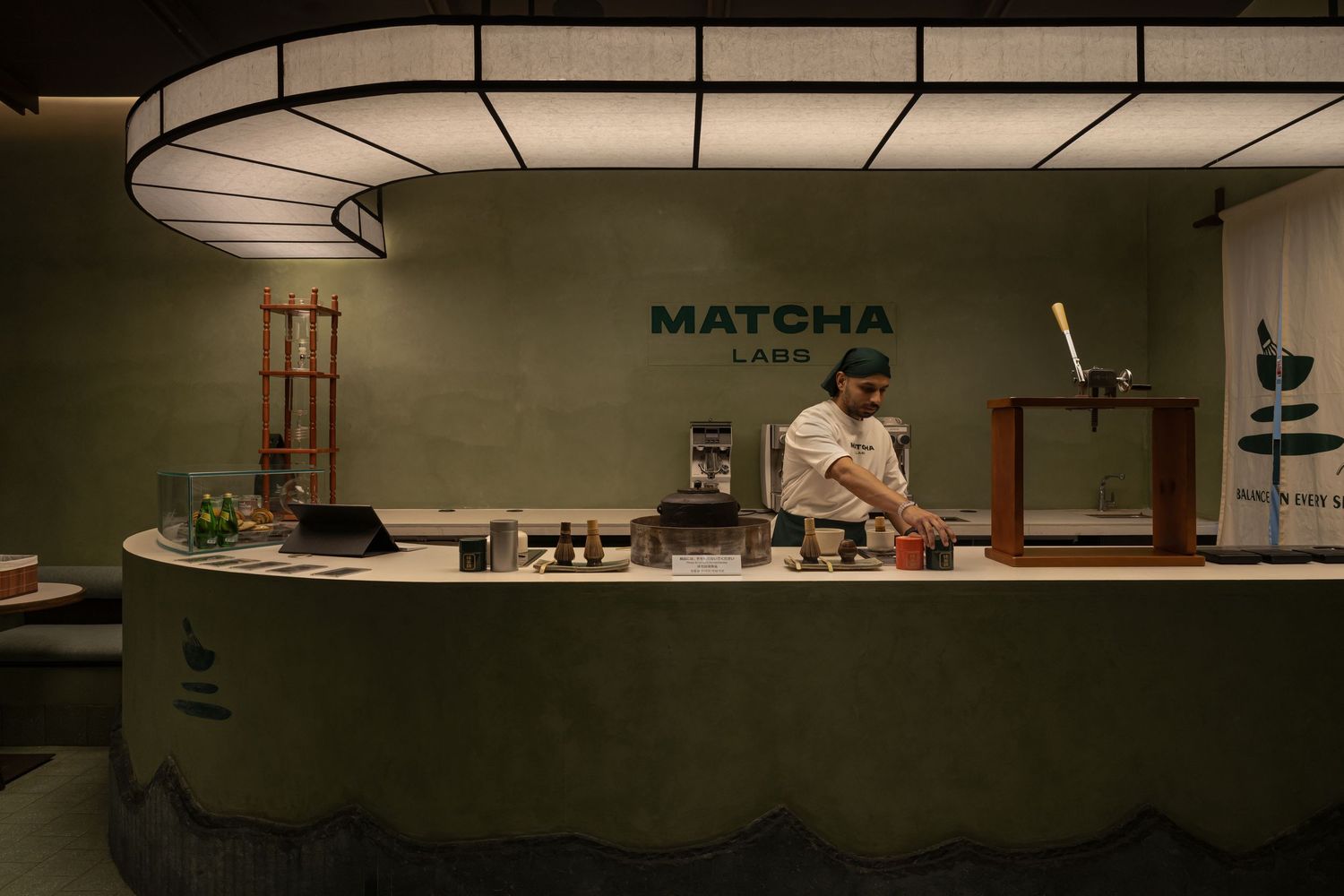 Matcha Labs cafe counter with curved green base, barista preparing drinks under soft oval pendant lighting.