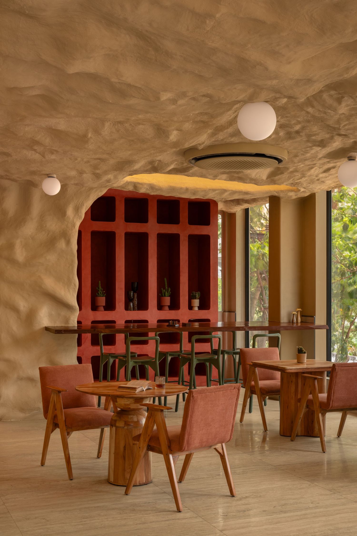 Restaurant interior with organic textured ceiling, red shelving unit, wooden tables, pink upholstered chairs, and globe ceiling lights.
