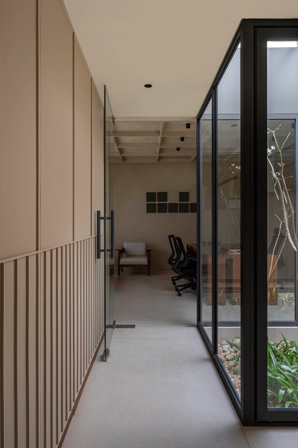Modern office corridor with warm beige walls, glass partition revealing indoor garden courtyard and workspace beyond.