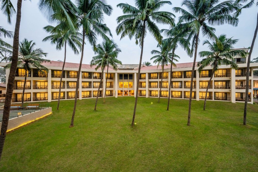 Tropical hotel exterior with curved architecture, lush green lawn, and tall swaying palm trees.