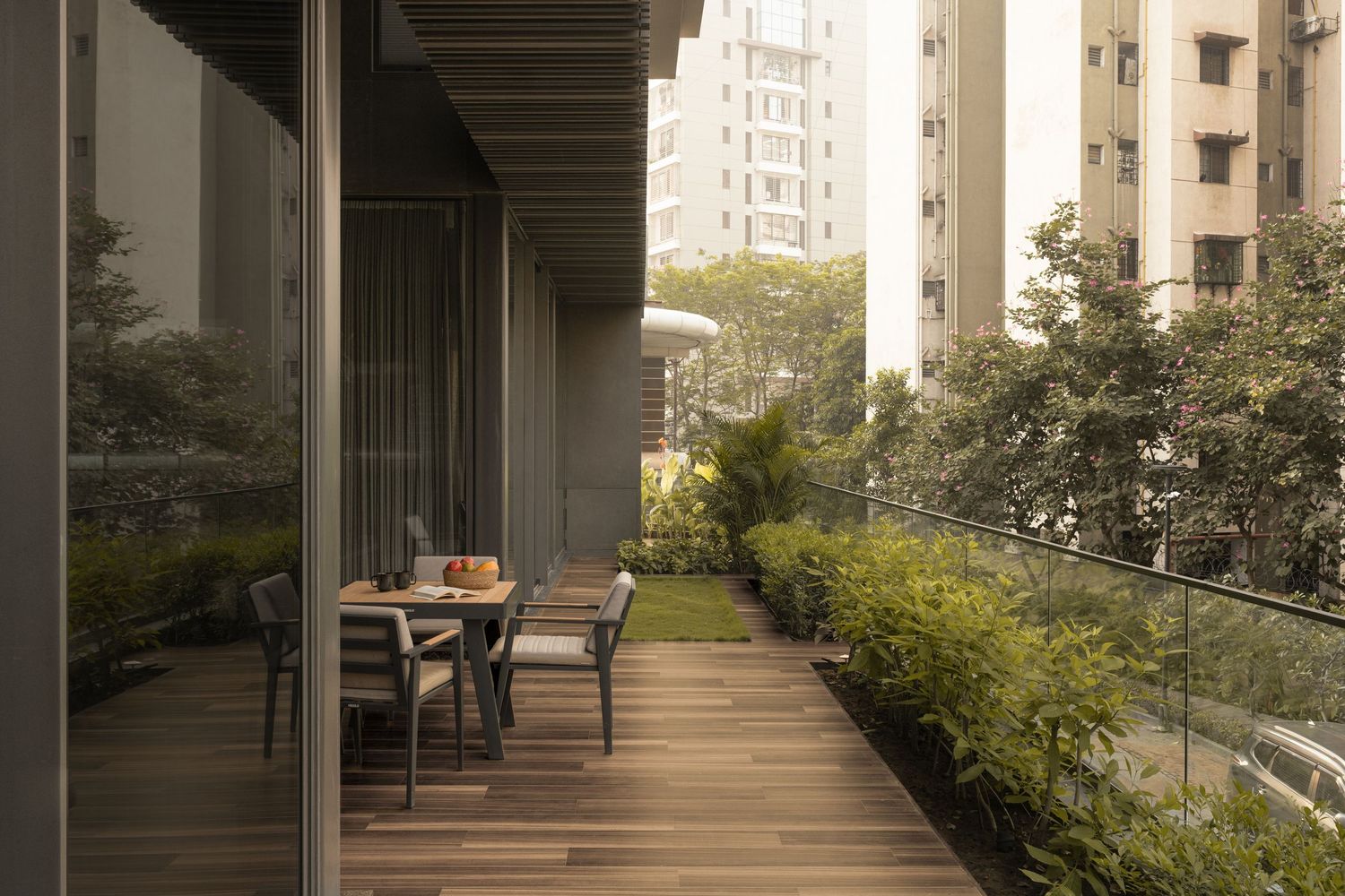 Elegant outdoor terrace with wooden deck, dining furniture, lush greenery, and glass railings overlooking city buildings.
