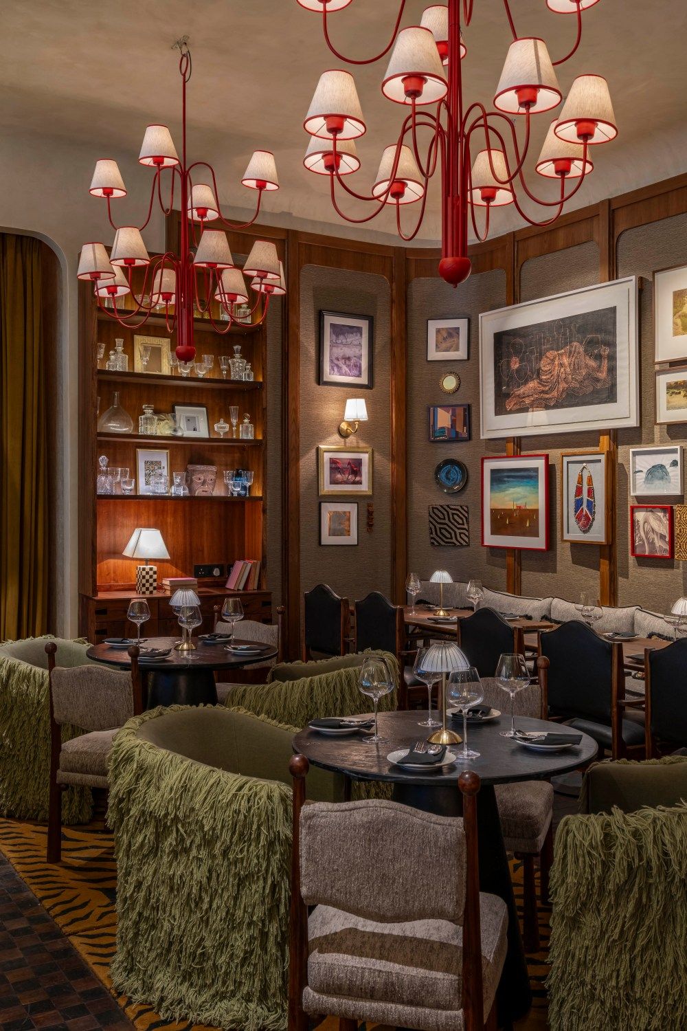 Warm restaurant interior with dramatic red chandelier, bookshelf display, green armchairs and gallery wall art.