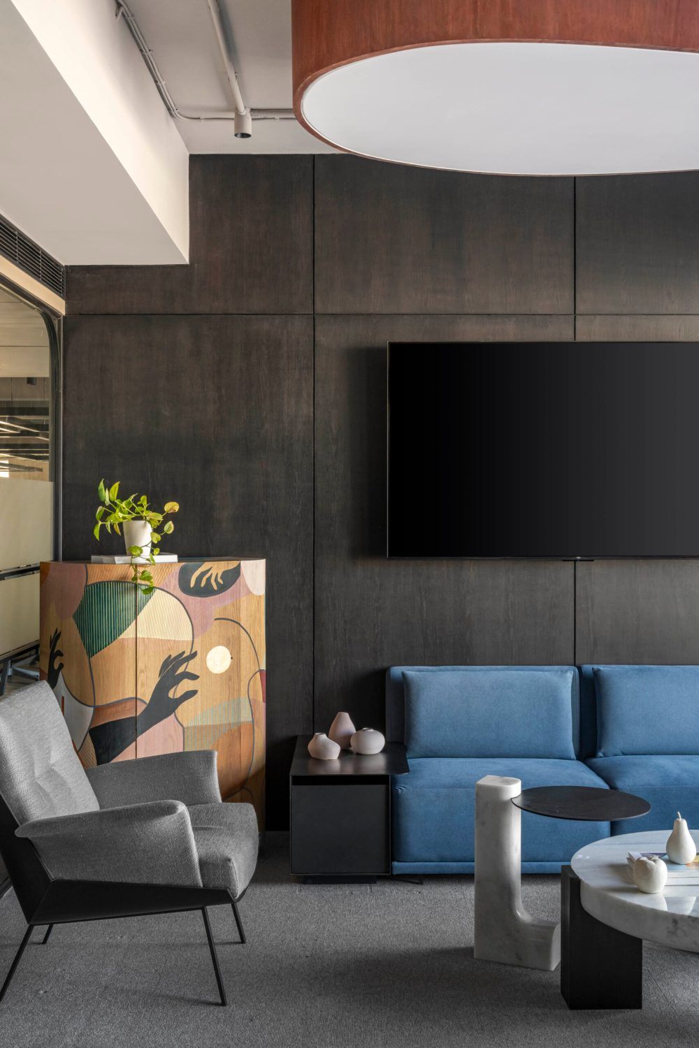 Modern office lounge with blue sofa, gray armchair, dark walls, and colorful artwork cabinet.
