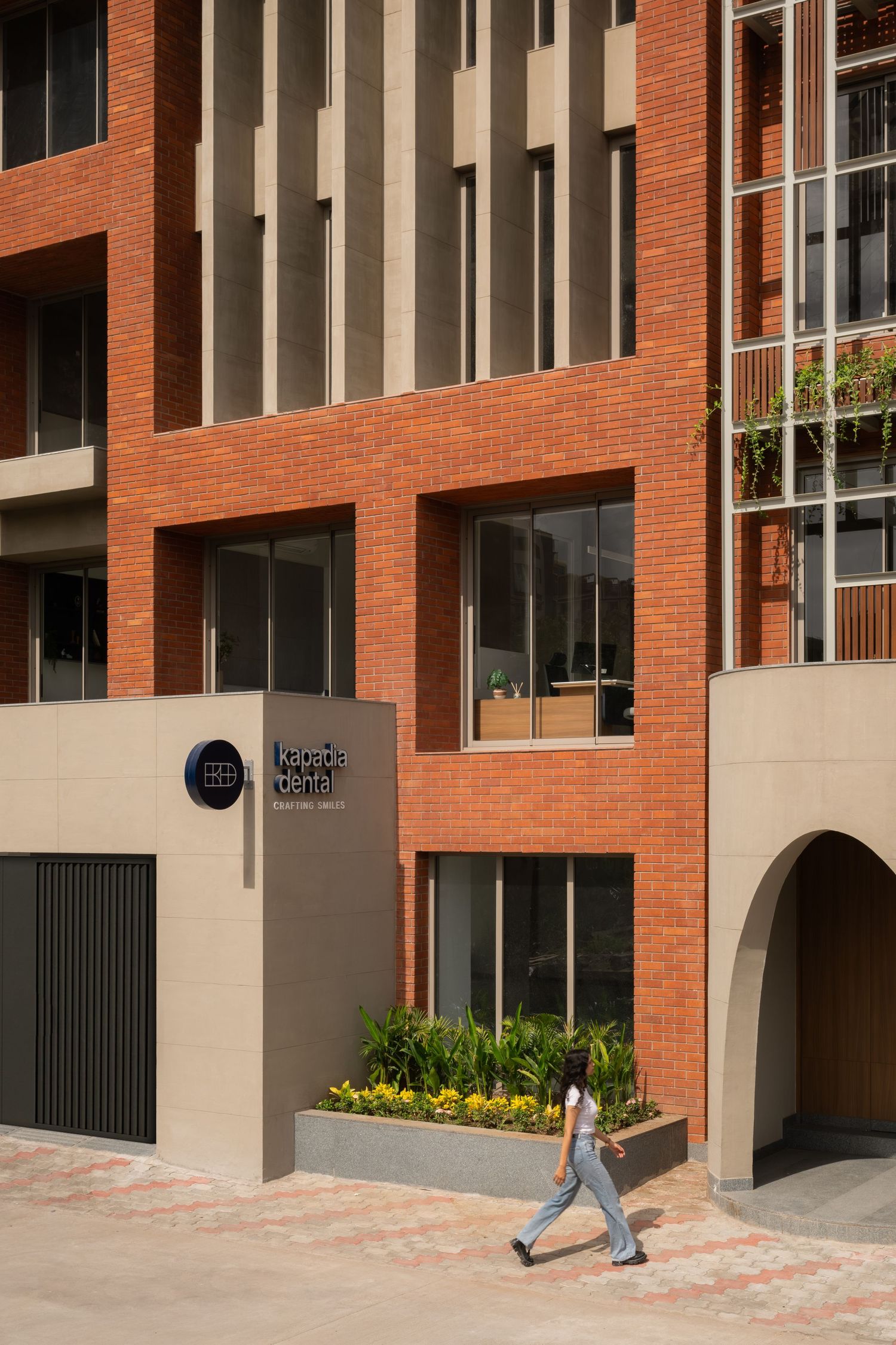 Close-up of red brick mixed-use building entrance with signage, yellow flowering plants, and pedestrian walking past arched facade.