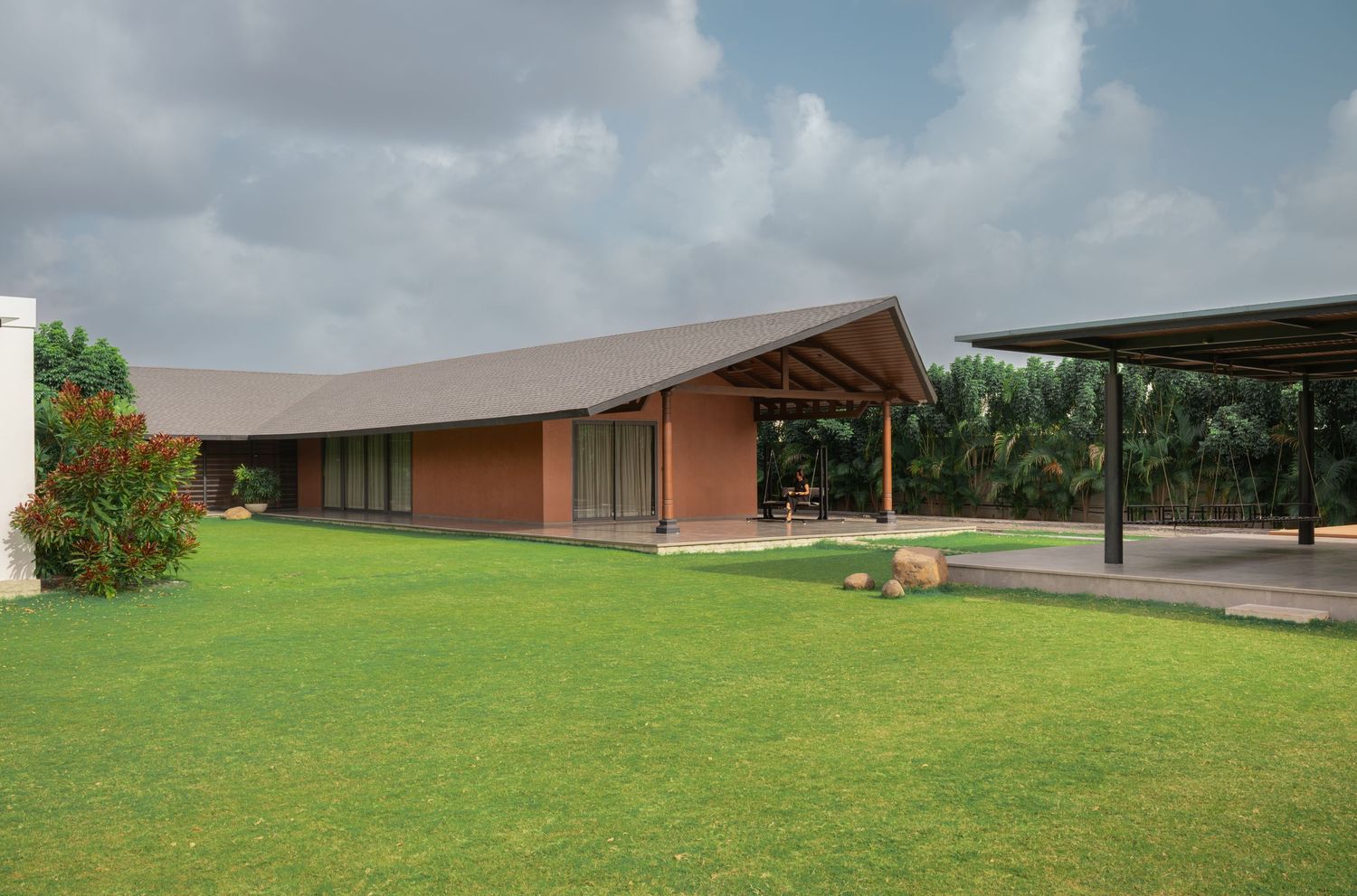 Low-profile terracotta residence with pitched roof, expansive green lawn, open pavilion structure, and tropical landscape backdrop.