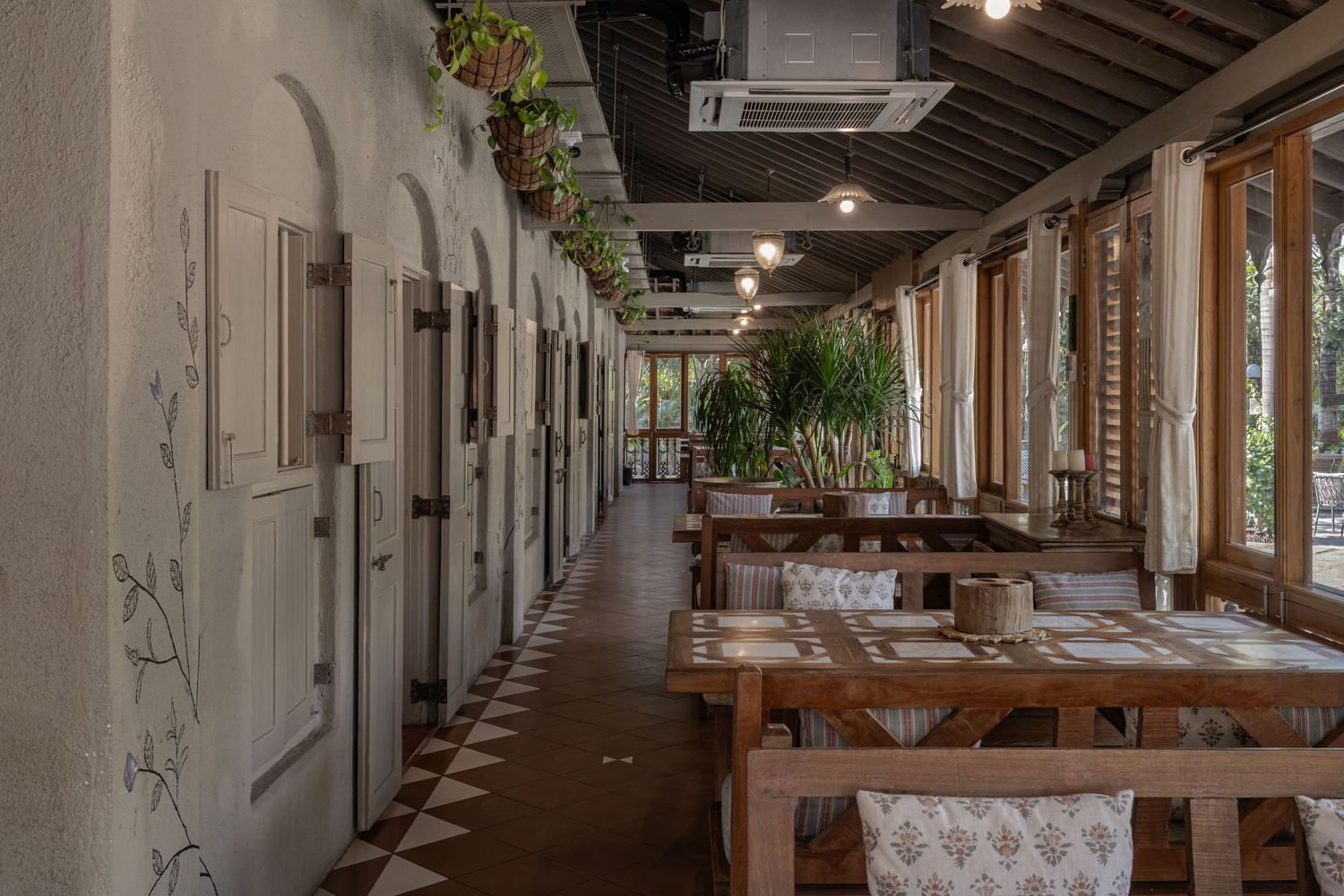 Rustic restaurant corridor with arched white walls, wooden tables, and lush hanging plants by large windows.