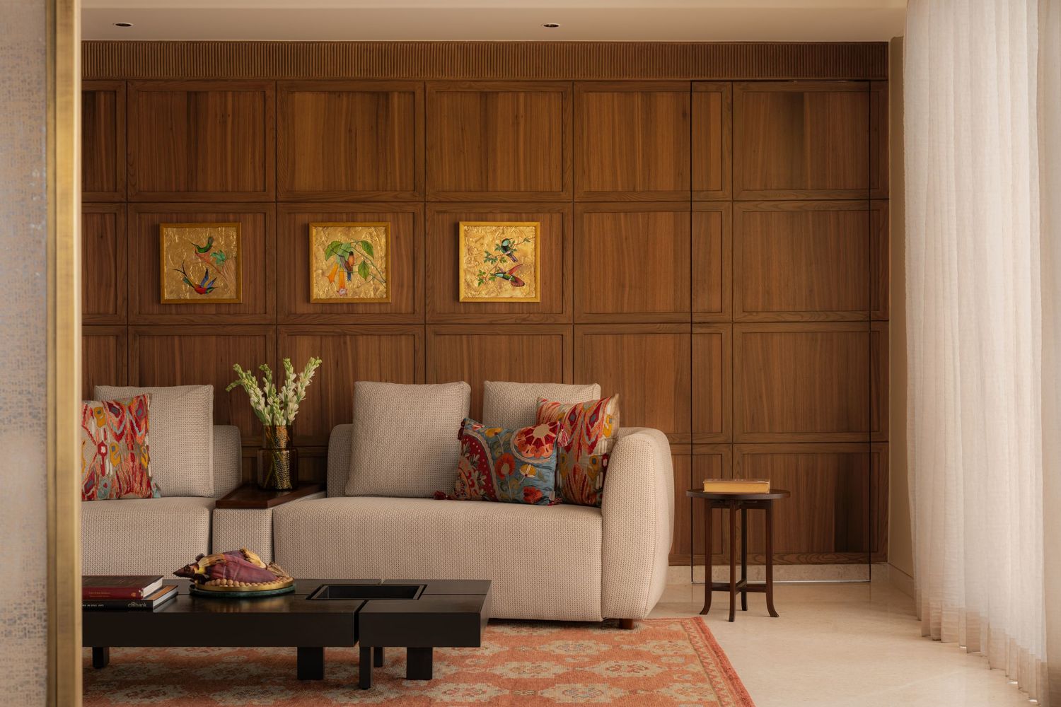 Warm living room with walnut wood panel wall, botanical artwork, white curved sofa, colorful cushions, and Persian rug.