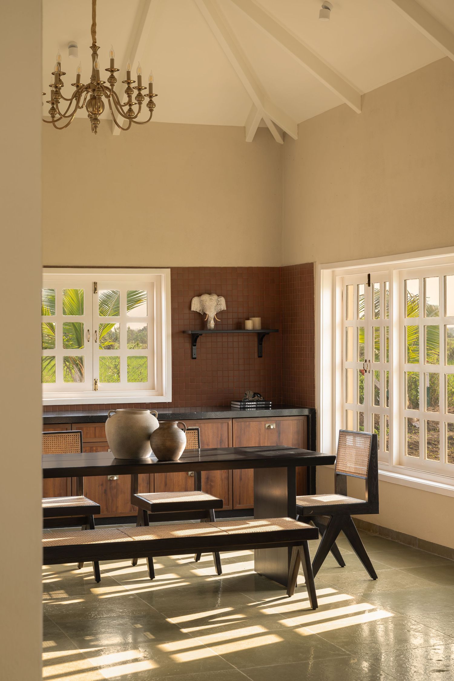 Rustic kitchen-dining area with vaulted ceiling, brass chandelier, wooden benches, ceramic vases, and garden views.