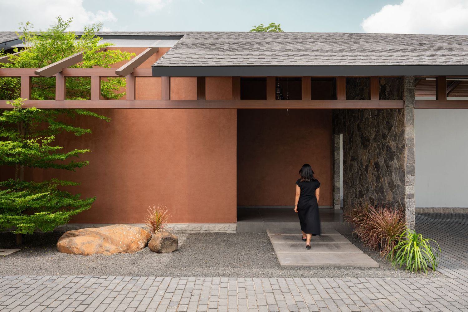 Terracotta walled home entrance with wooden pergola, natural stone boulder, lush greenery, and woman walking toward doorway.
