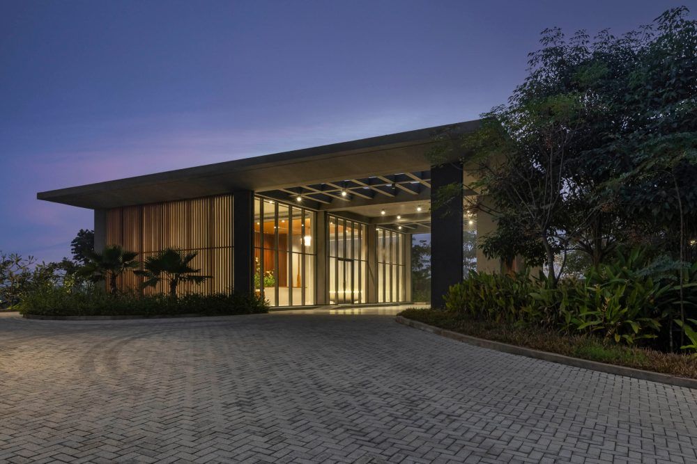 Illuminated modern building entrance at dusk with flat roof, glass facade, stone pavers, and surrounding tropical landscaping.