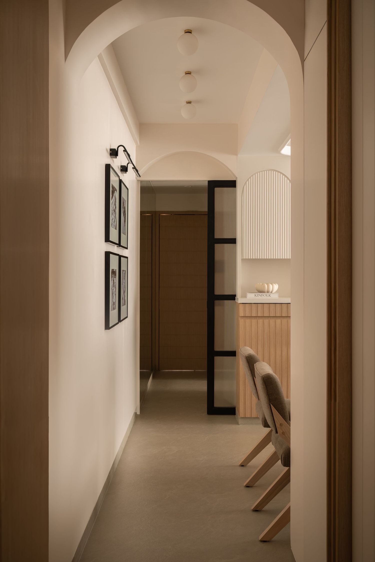 Modern hallway with arched ceilings, framed wall art, wooden door, and dining chairs in a minimalist neutral interior.