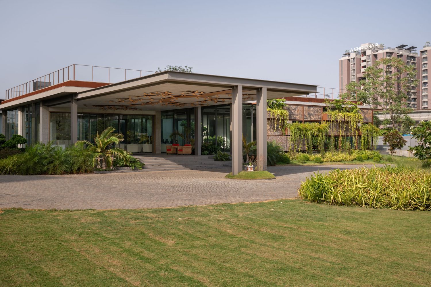 Modern pavilion building with open facade, lush green lawn, tropical plants, and high-rise apartments in background.