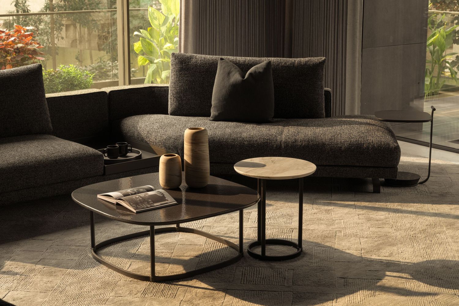 Contemporary living room with dark gray sofa, nesting coffee tables, and sunlight streaming through large garden windows.
