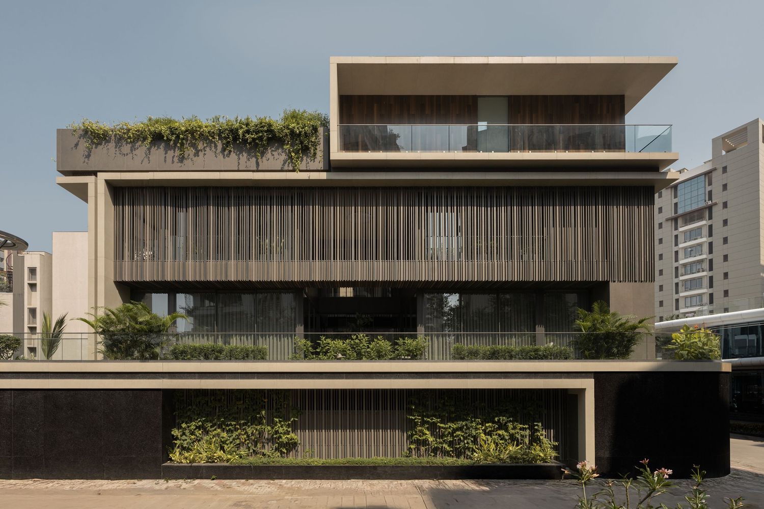 Modern three-story building with vertical wood louvers, lush green planters, and rooftop garden in daylight.