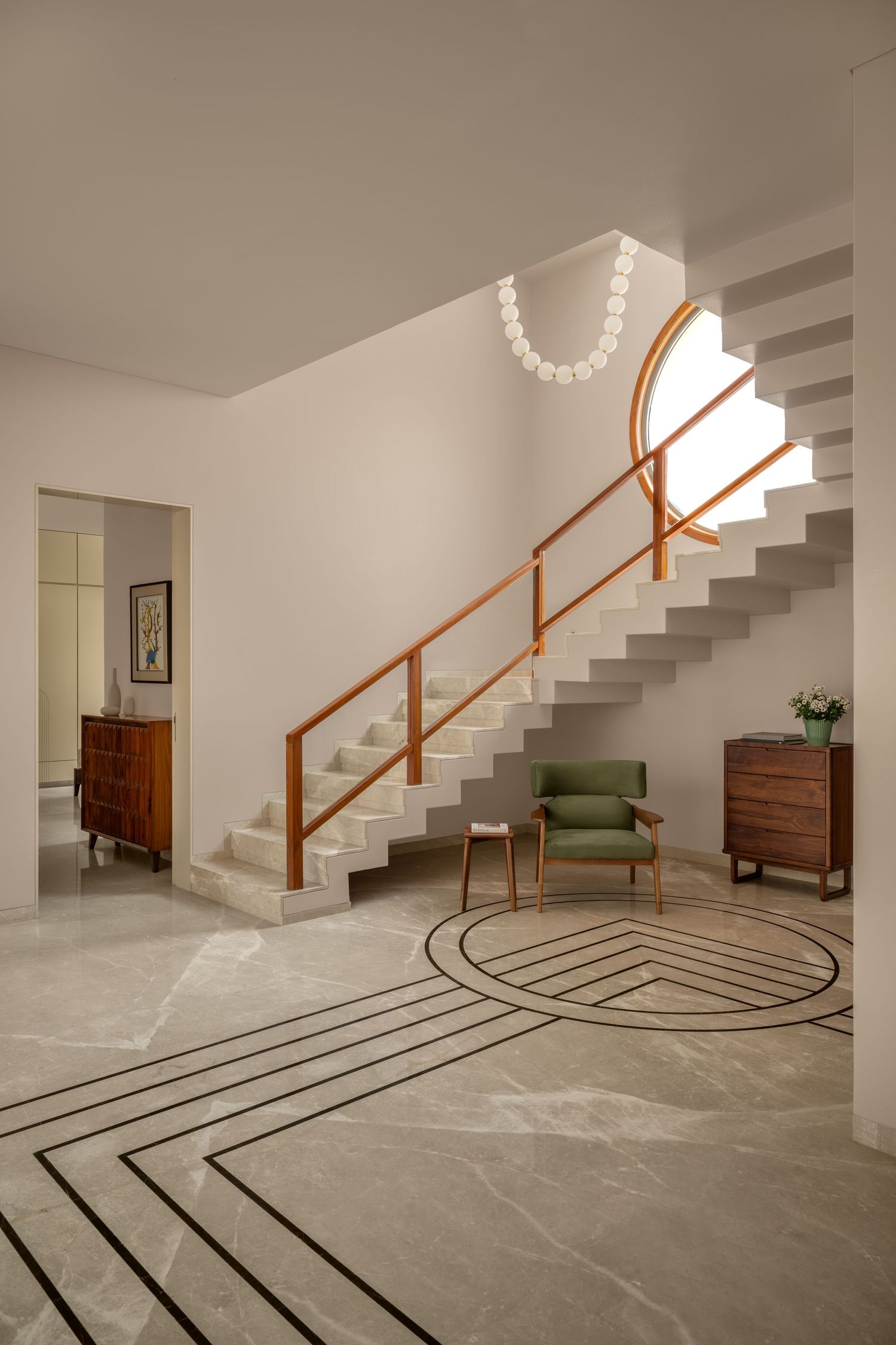 Minimalist foyer with white staircase, wooden handrail, green accent chair, marble floor inlay, and wooden dressers.