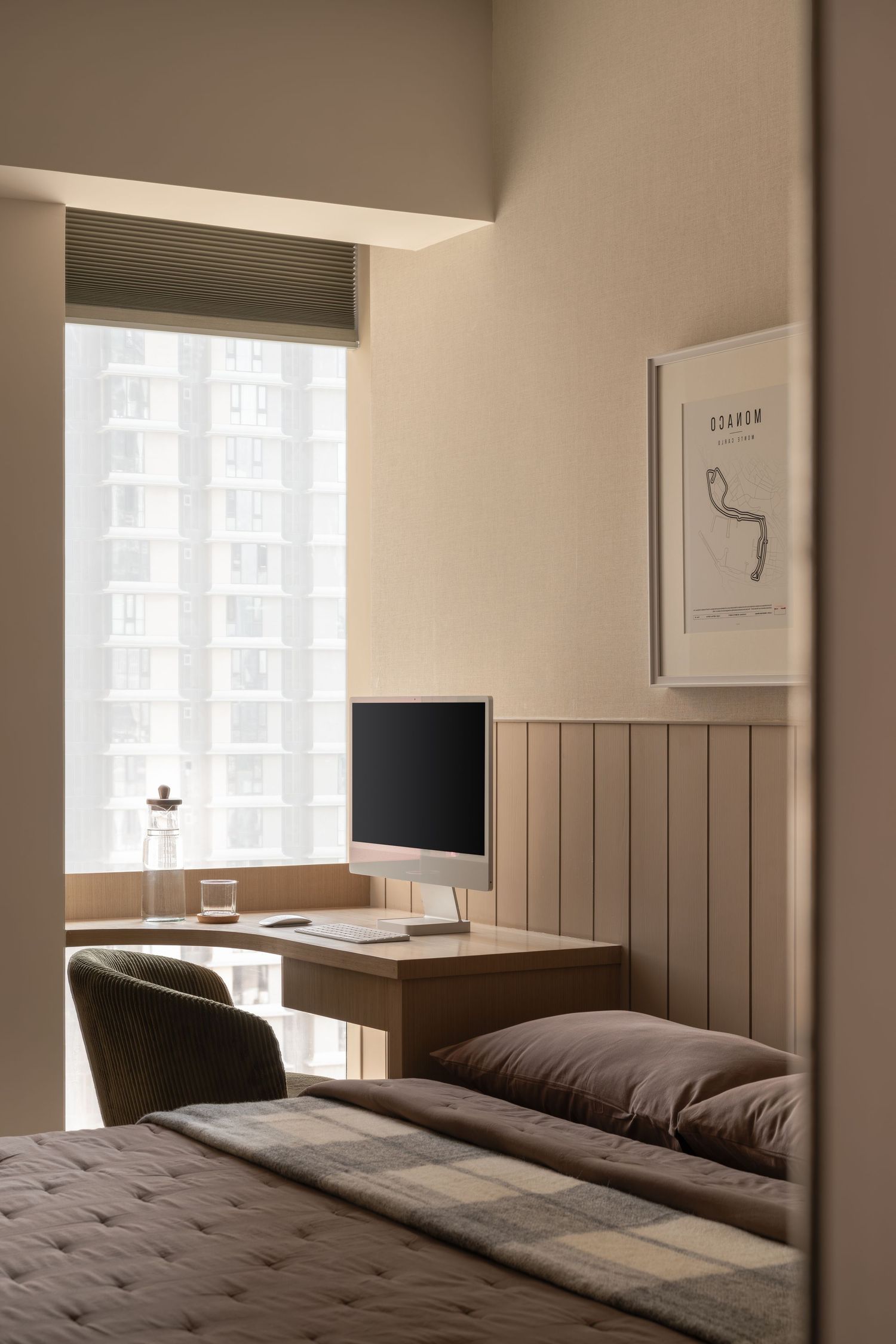 Minimalist bedroom with built-in desk, computer monitor, and large window overlooking city buildings.