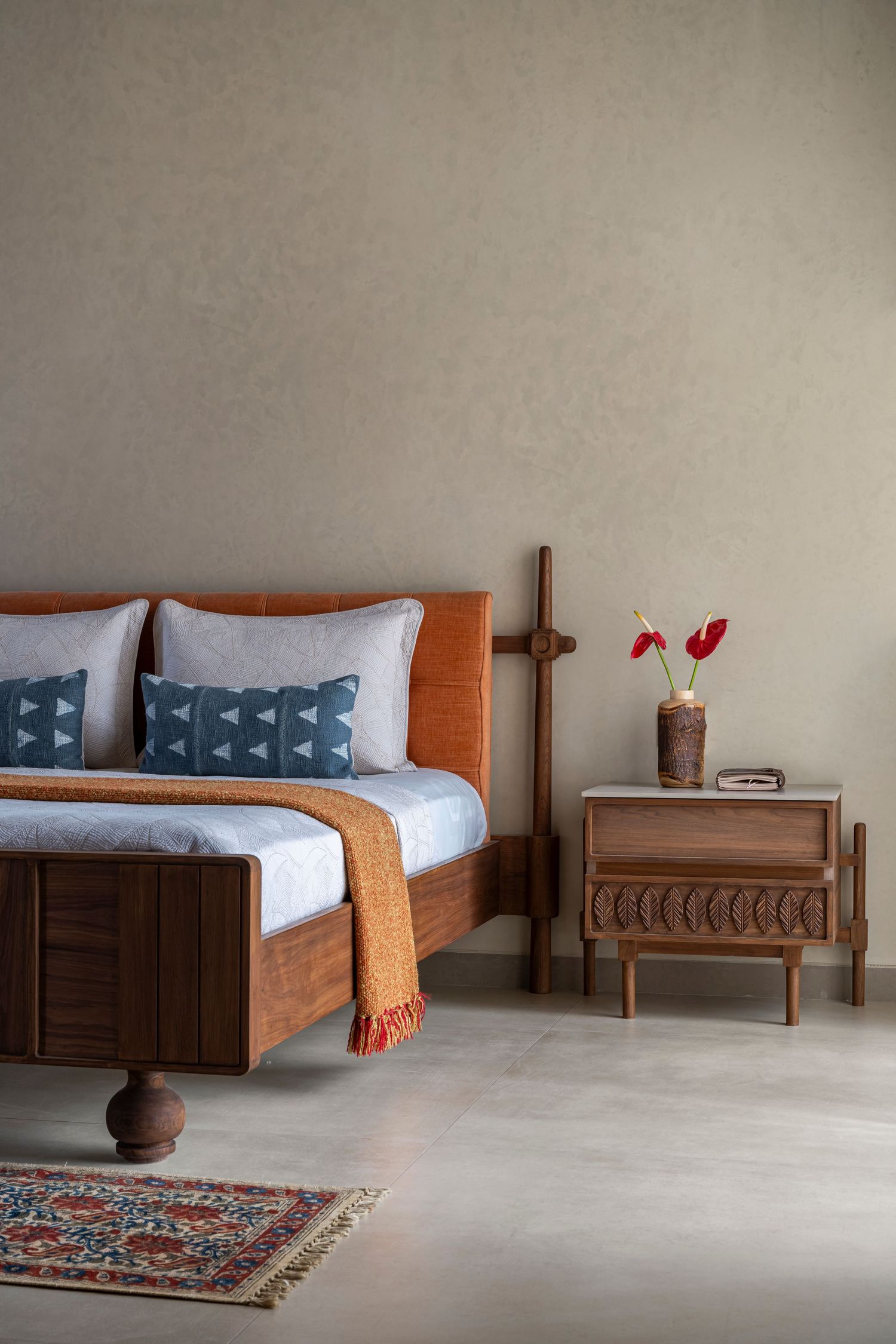Elegant bedroom with wooden bed frame, orange throw, patterned rug, and nightstand with red flowers on beige wall.