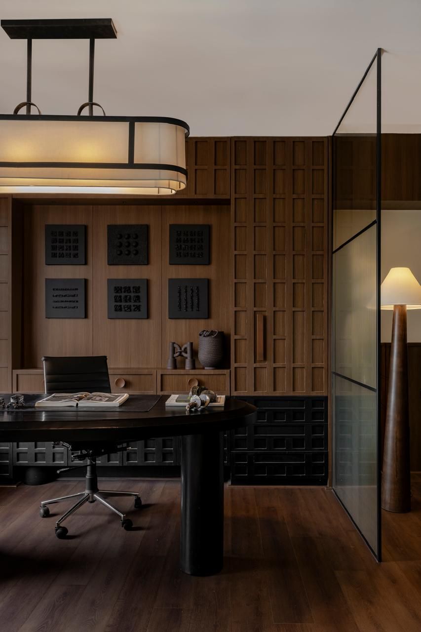 Dark wood executive office with grid wall panels, oval desk, ergonomic chair, and warm pendant lighting.