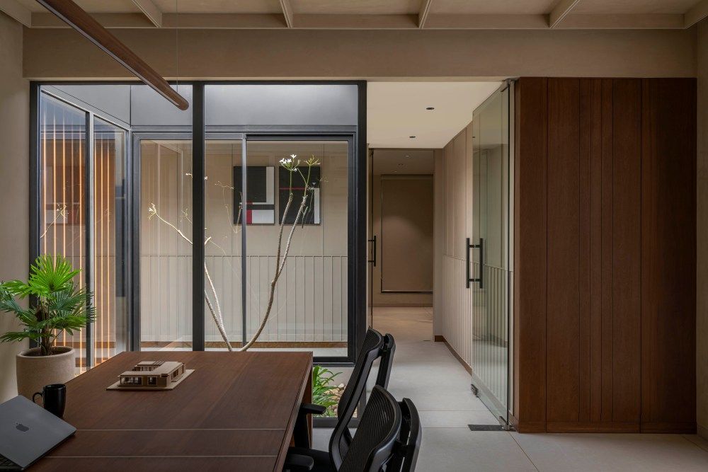 Modern office interior with glass partition walls, wooden dining table, black chairs, and indoor garden courtyard view.
