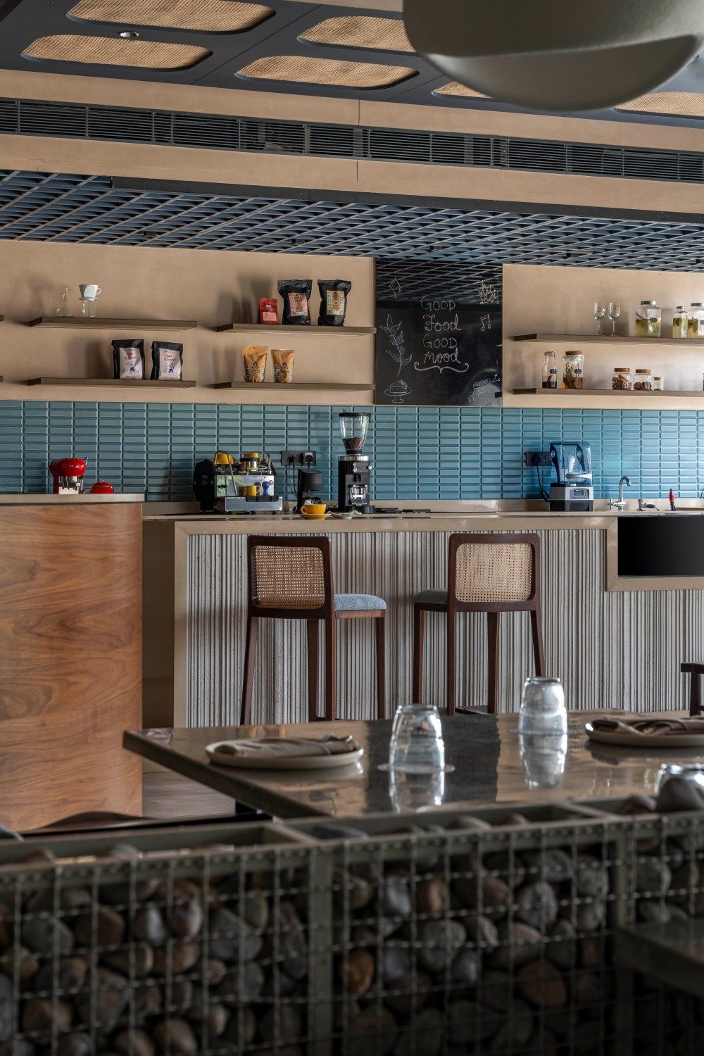 Close-up of restaurant bar station with blue tile wall, wooden shelving, rattan chairs, and coffee equipment on counter.