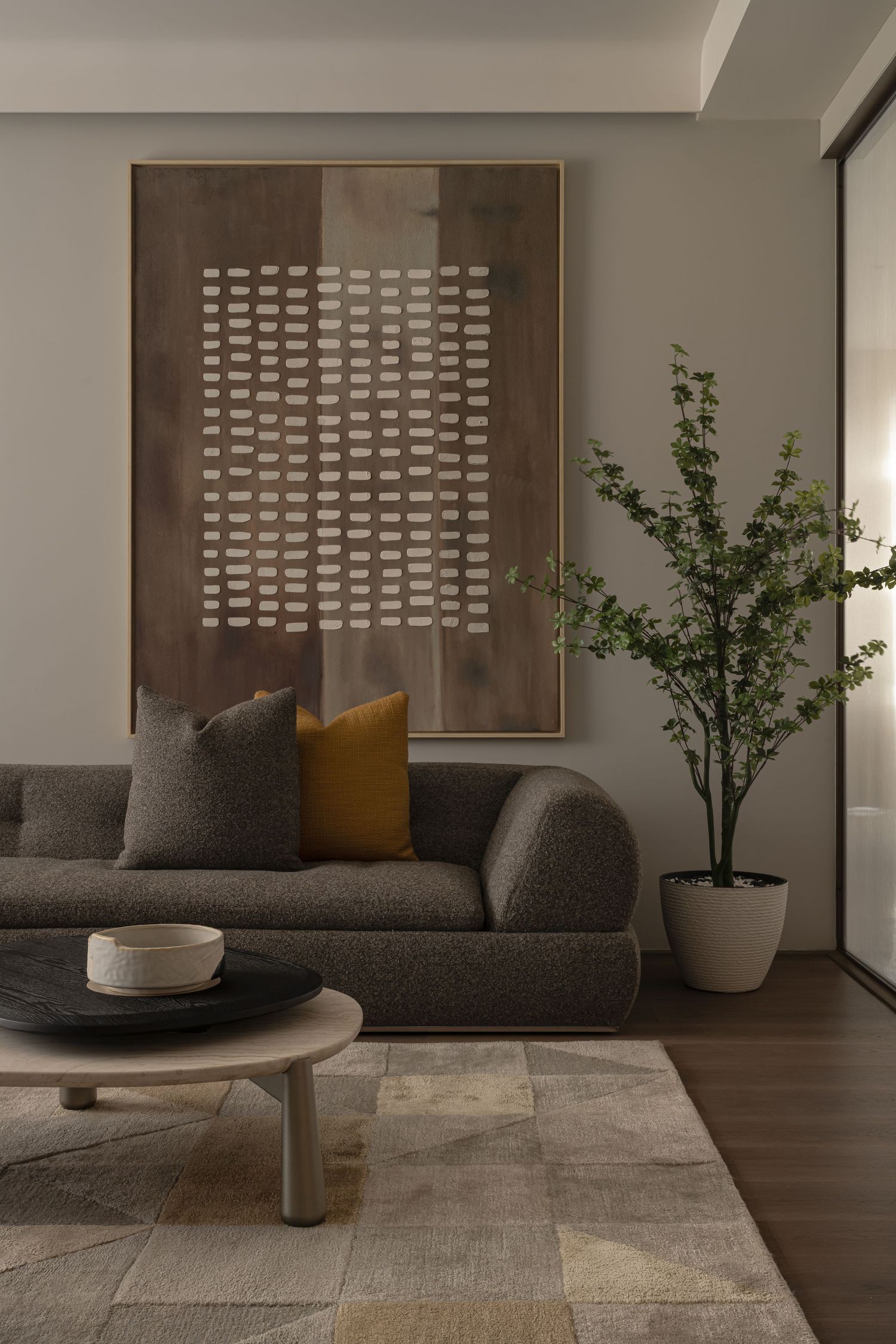 Cozy living room with gray boucle sofa, mustard pillow, abstract brown artwork, and potted tree.