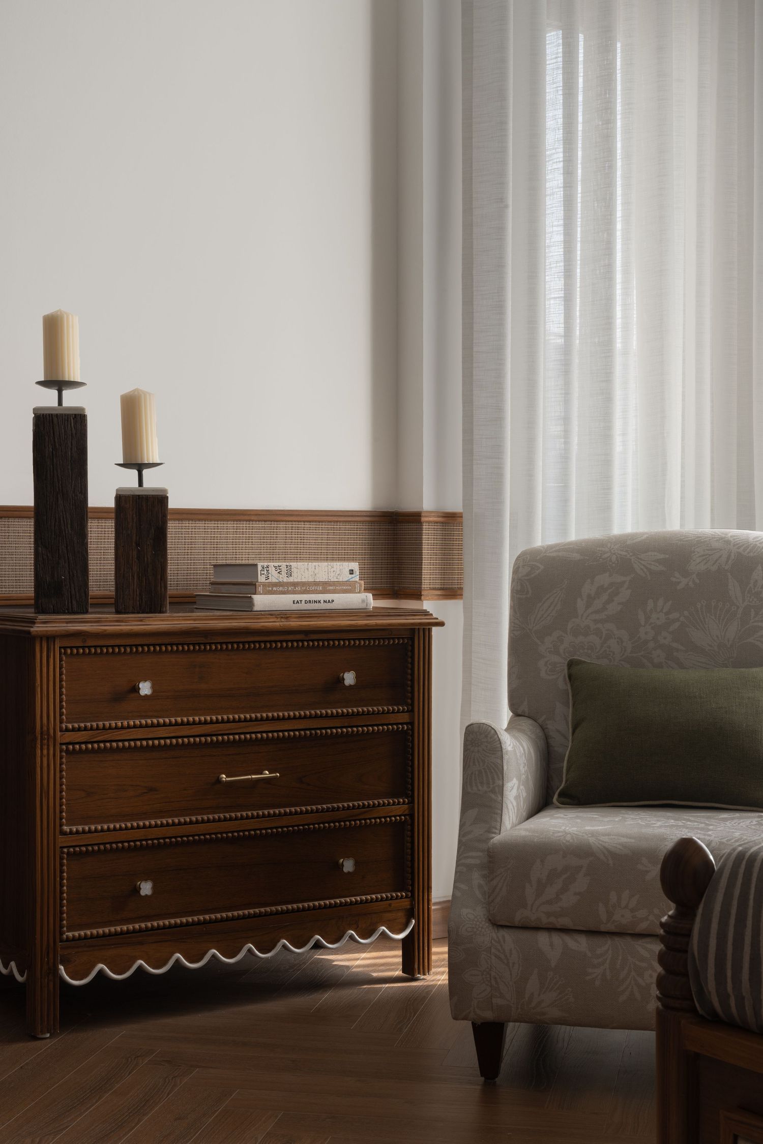 Classic wooden dresser with candle holders beside a floral patterned armchair near sheer curtained window.