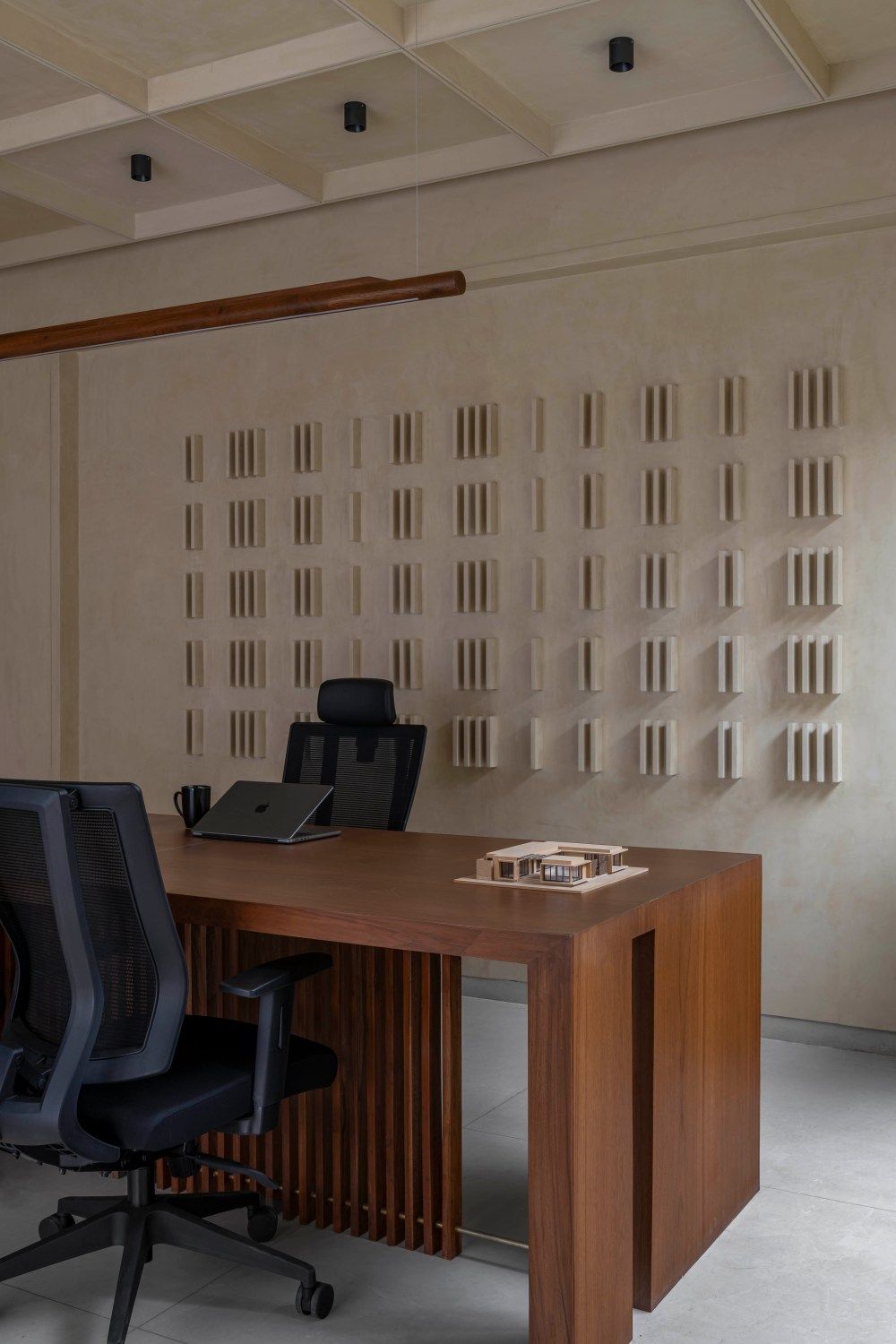 Minimalist office workspace with walnut wood slatted desk, black ergonomic chairs, laptop, and textured decorative wall panels.