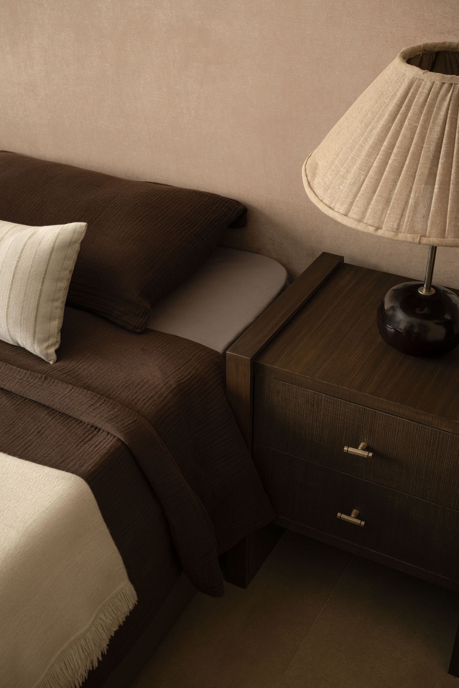 Close-up of dark brown bedroom with textured bedding, striped pillow, and pleated table lamp on nightstand.