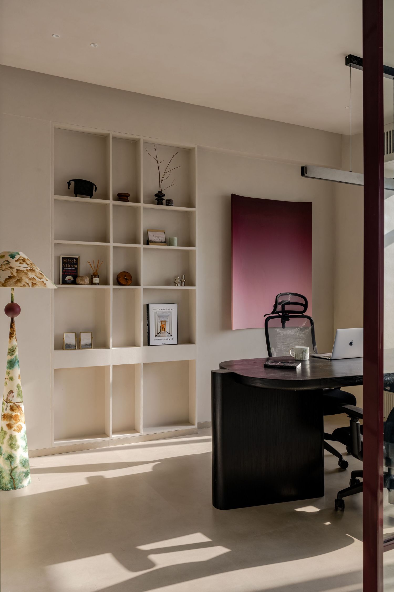 Minimalist home office with dark curved desk, ergonomic chair, cream shelving, and pink abstract wall art.