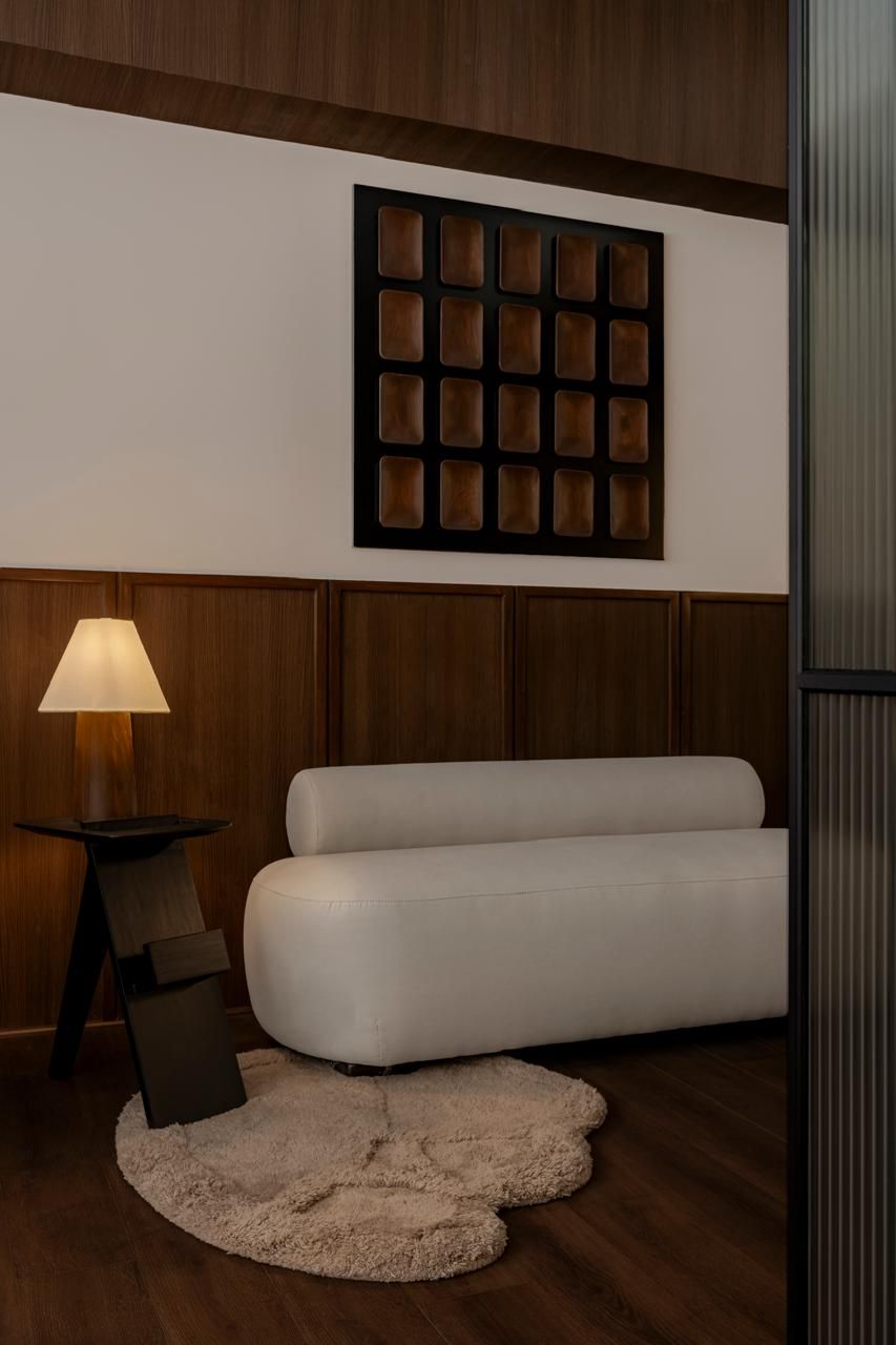 Modern lounge with white curved sofa, fluffy rug, grid wall art, and warm lamp on dark wood paneling.