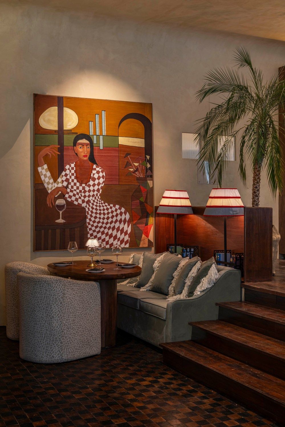 Cozy restaurant lounge with large figurative painting, gray sofa, tall lamps, and tropical palm tree decor.