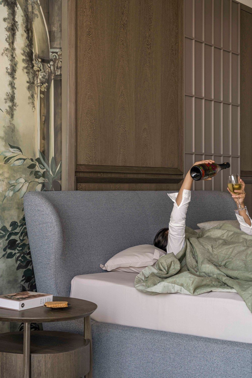 Person lying in gray upholstered bed pouring wine, with botanical mural wallpaper and wooden slat wall panel.