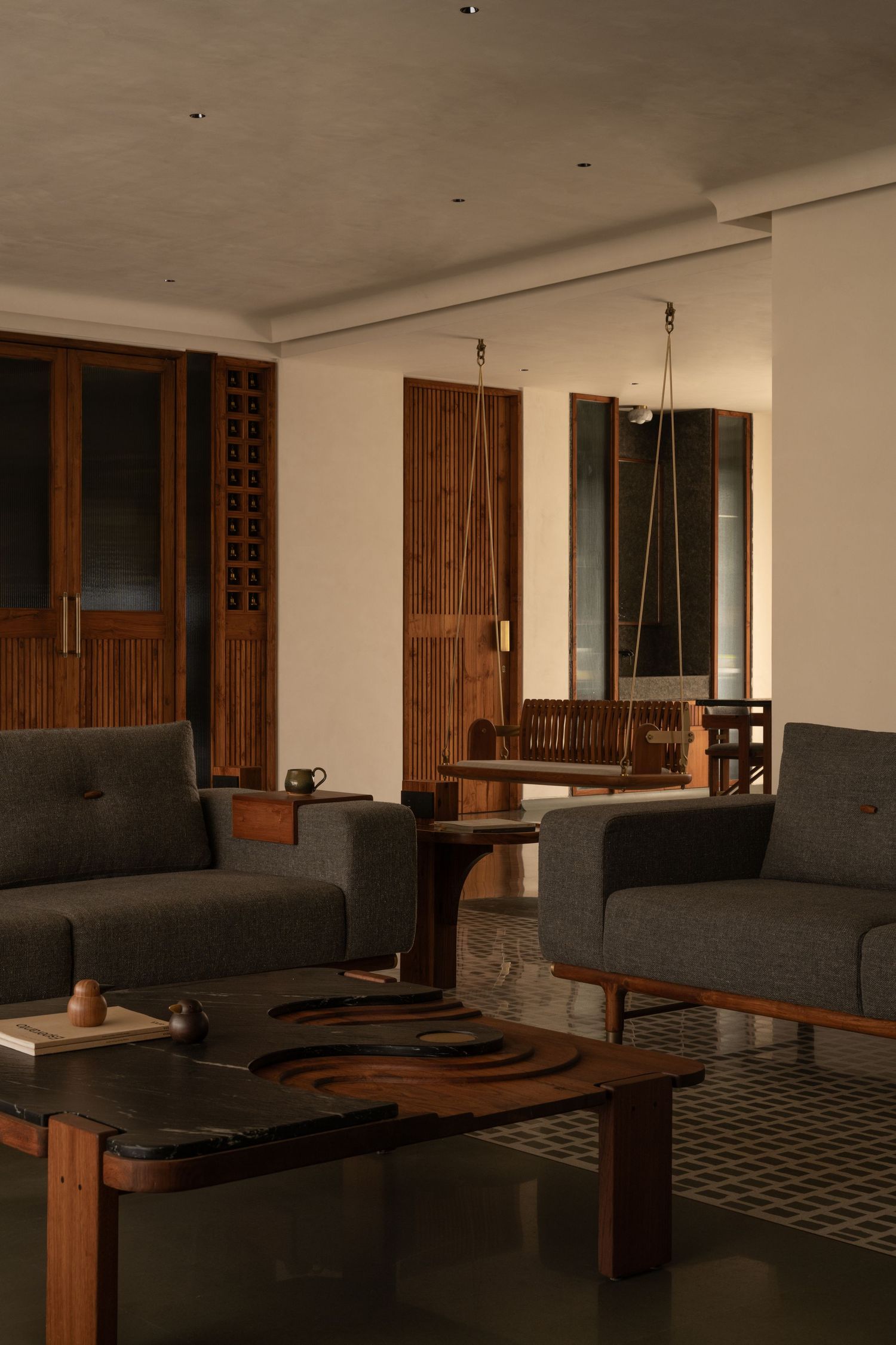 Dimly lit living room with gray sofas, wooden swing, lattice doors, and patterned tile floors.