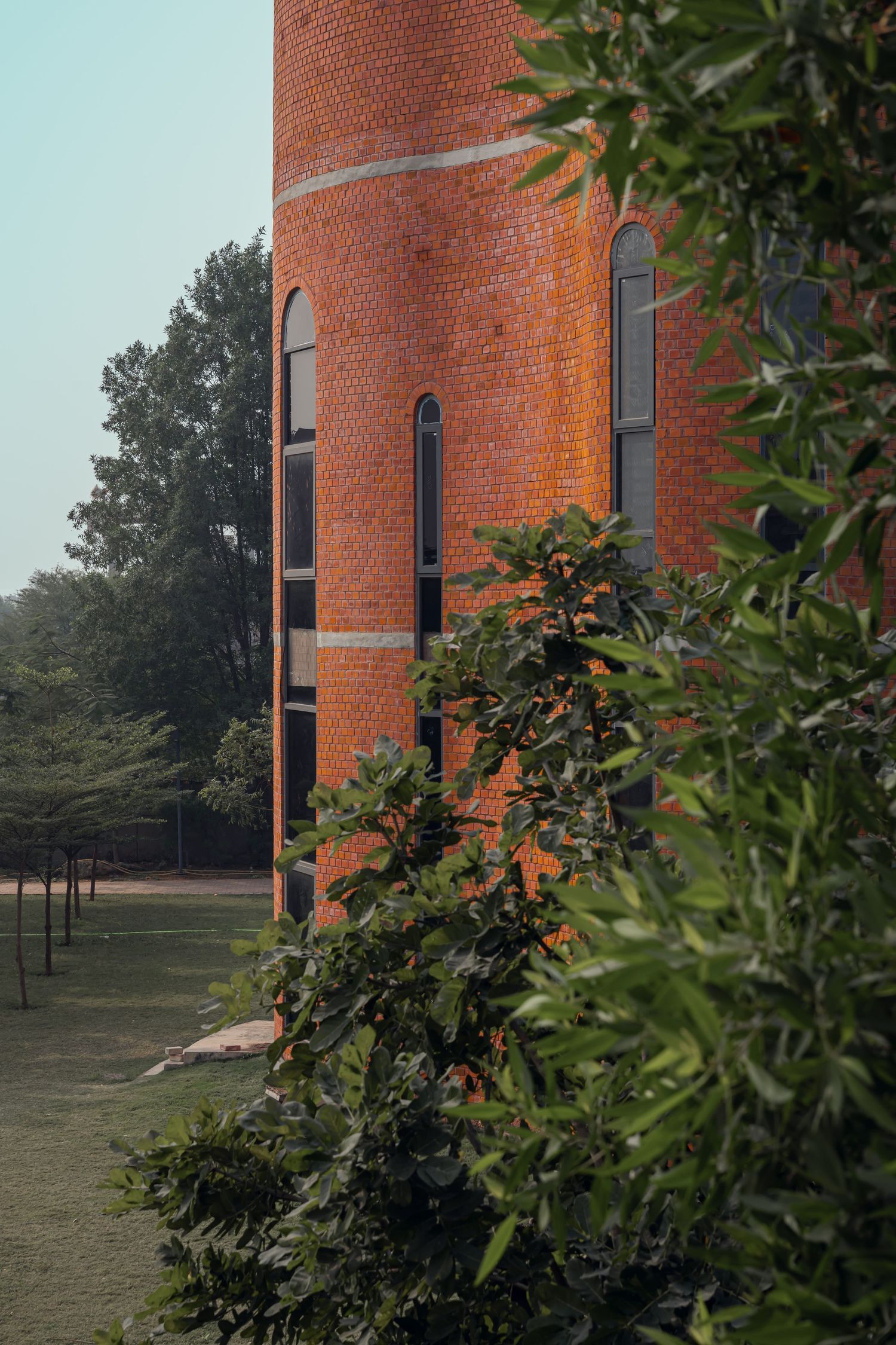 Tall red brick cylindrical tower with arched windows surrounded by lush green trees.
