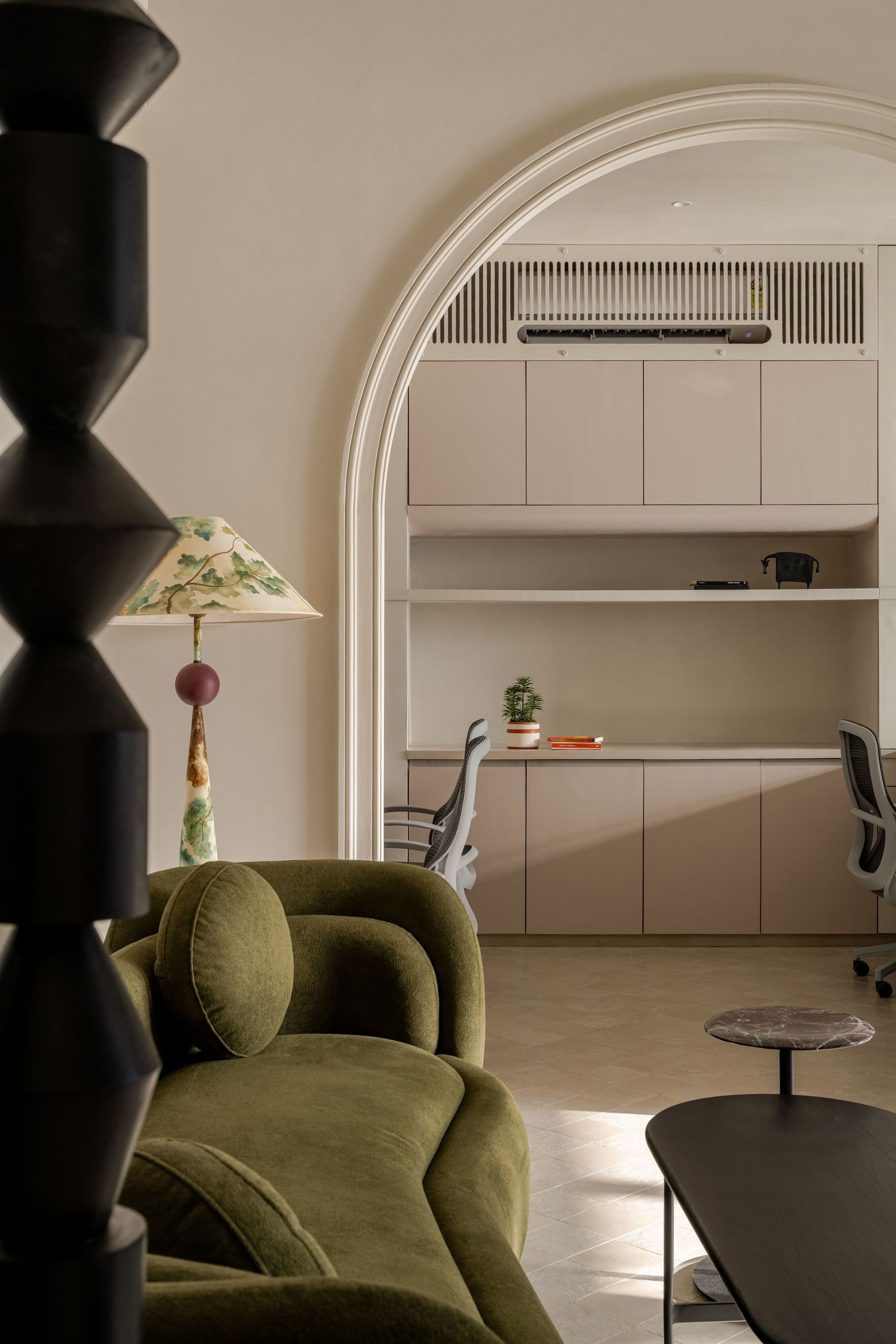 Cozy interior with green velvet sofa, decorative floral floor lamp, arched doorway to beige home office workspace.